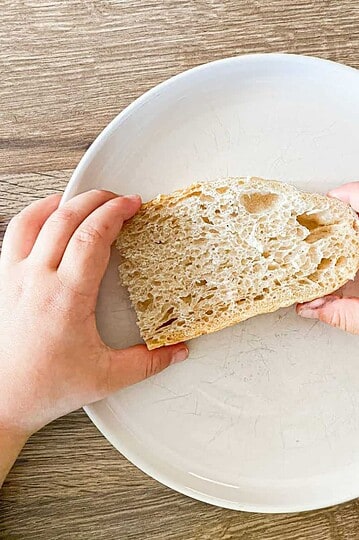 Child holding a piece of soft sourdough bread on a white plate.