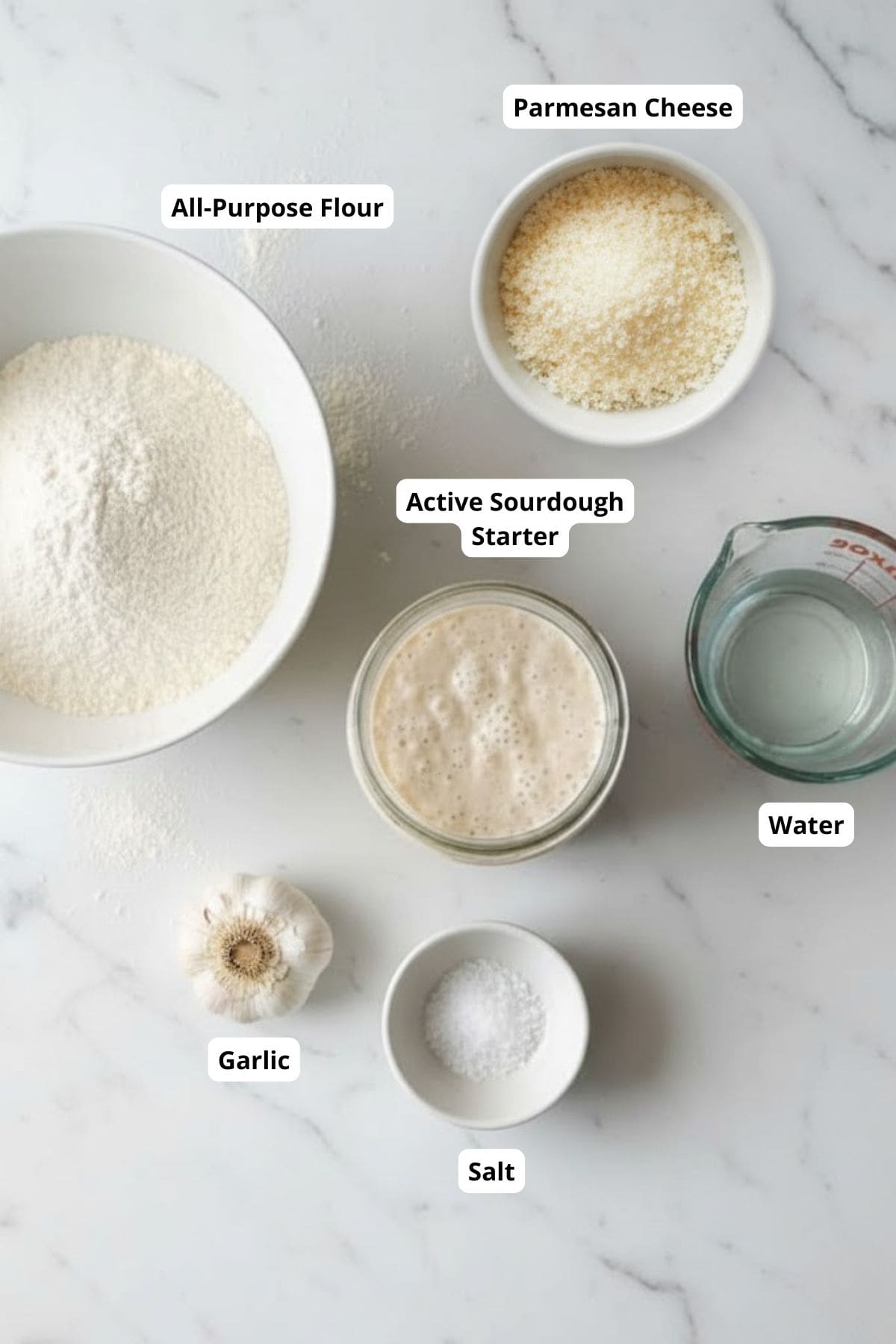 All-purpose flour, active sourdough starter, water, garlic, parmesan cheese, and salt on a white marble surface.