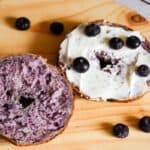 Sourdough Blueberry bagel cut in half, greek yogurt spread over one half, on a round wooden board.