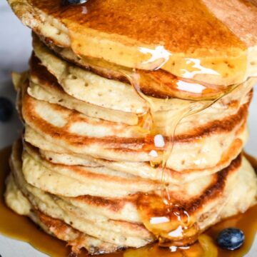 Sourdough banana pancakes stacked up on a plate, with maple syrup and blueberries.