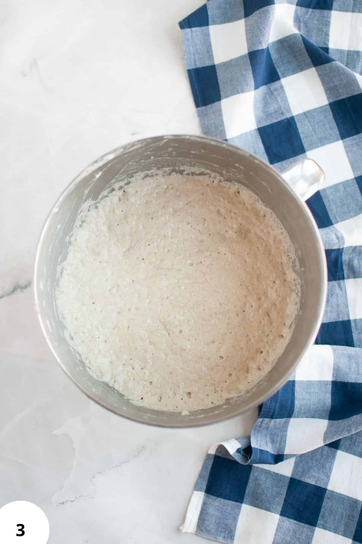 Dough in a mixing bowl ready for stretch and folds, on a marble surface with a blue checkered cloth.
