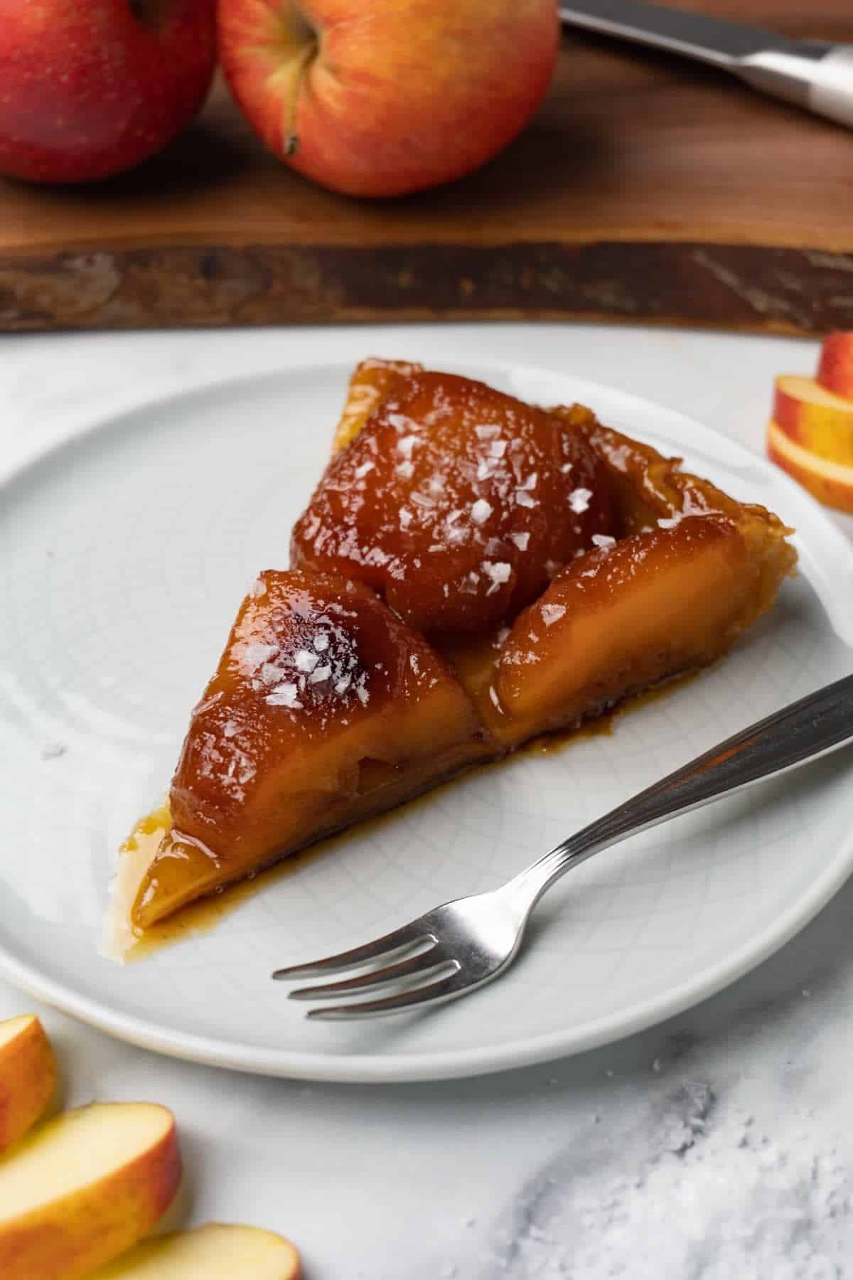 A single slice of apple tarte tatin on a plate, served with a fork on the side.