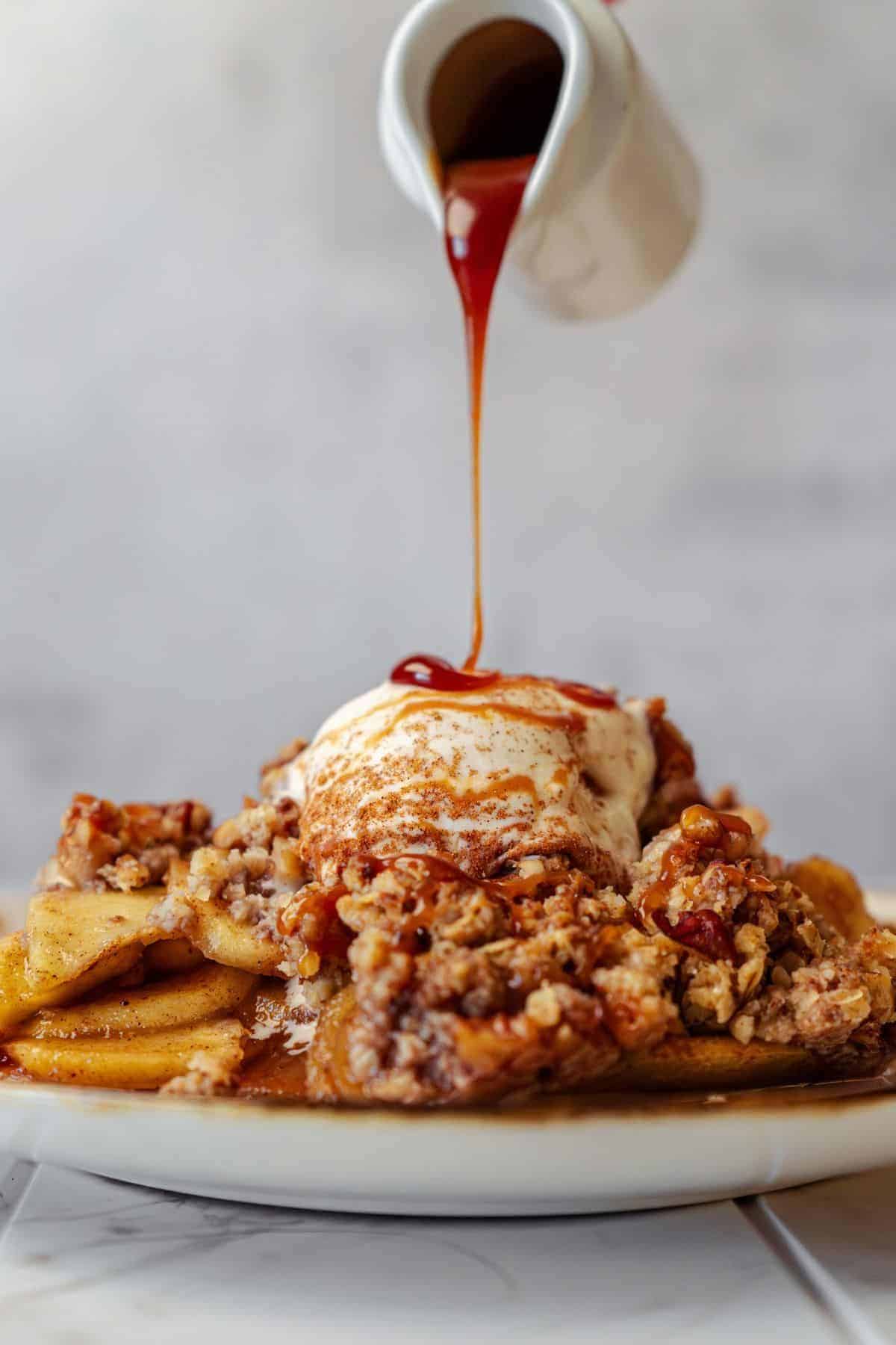 A serving plate of warm apple crisp topped with a scoop of vanilla ice cream and drizzled with caramel sauce.