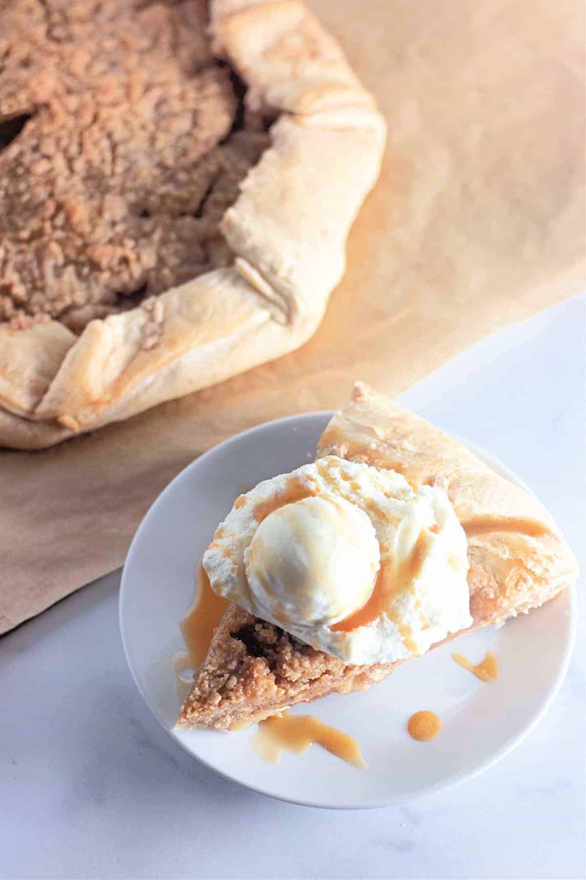 Slice of apple galette on a plate topped with vanilla ice cream, with the remaining whole galette in the background.