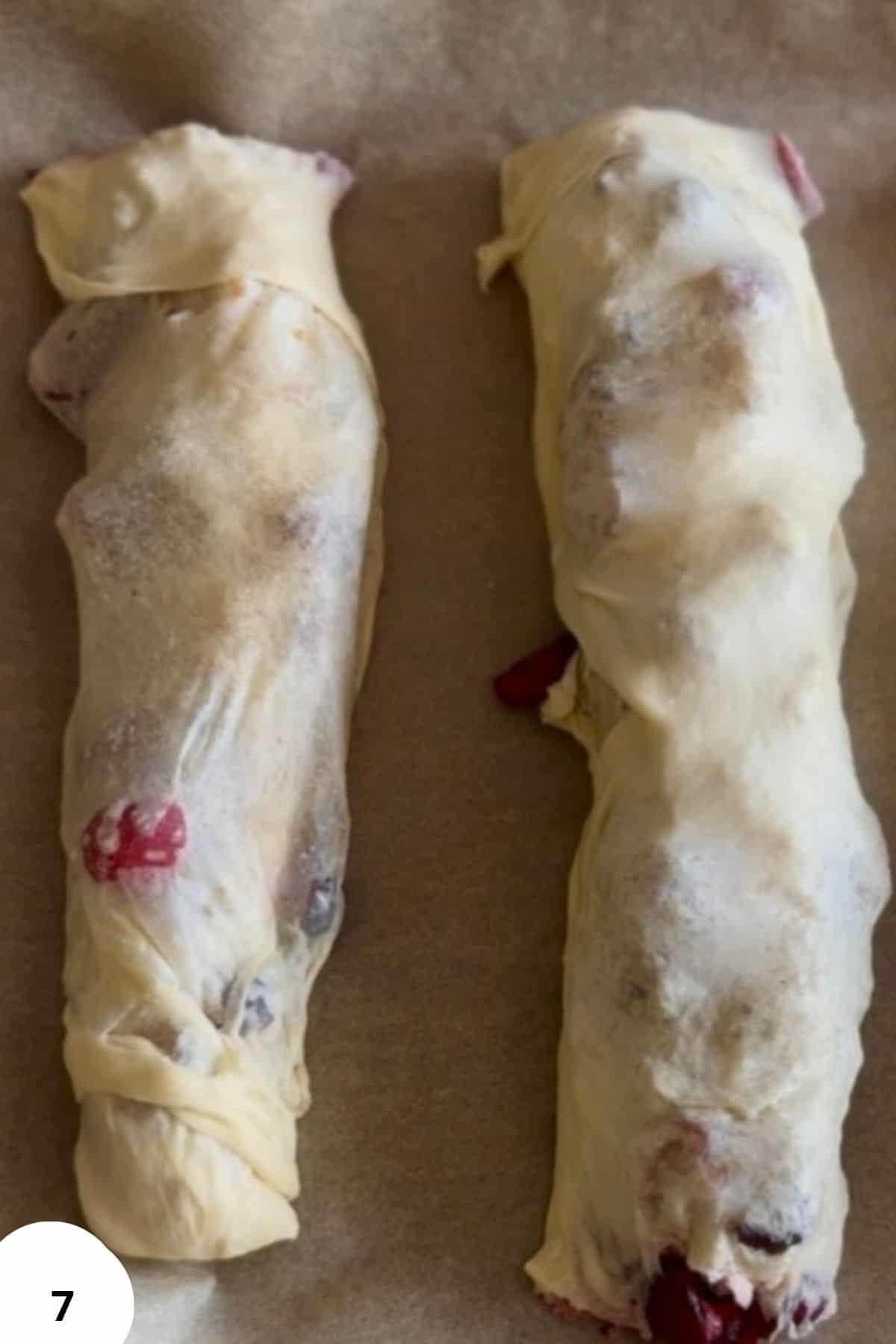 Two unbaked cherry strudel logs placed on a parchment-lined baking sheet, ready for the oven.