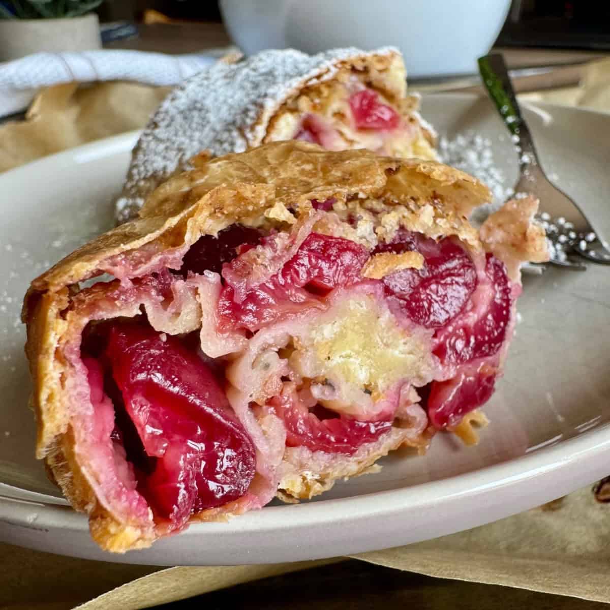 A slice of cherry strudel served on a plate, showing the interior layers of cherries, breadcrumbs, and flaky pastry.