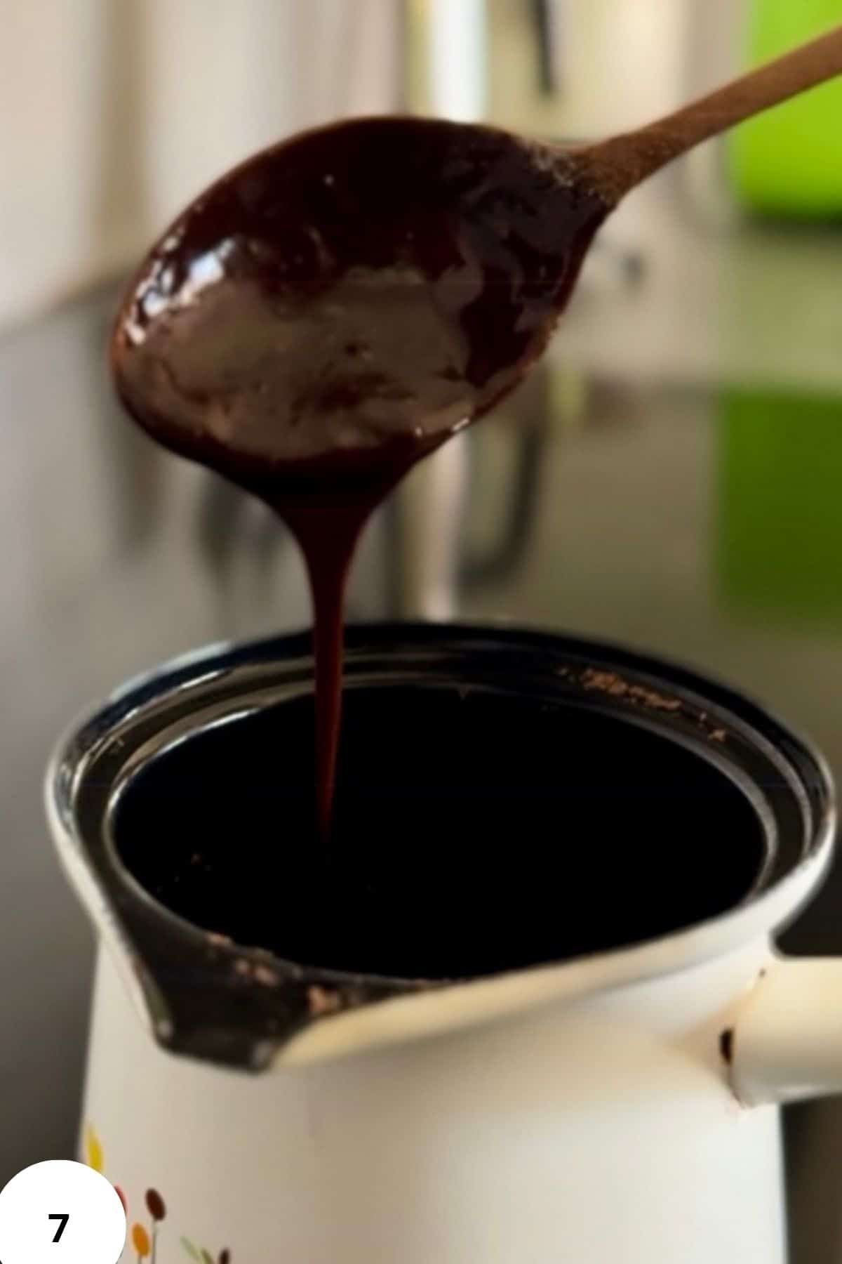 A spoon pouring melted chocolate from a small pitcher-style saucepan.