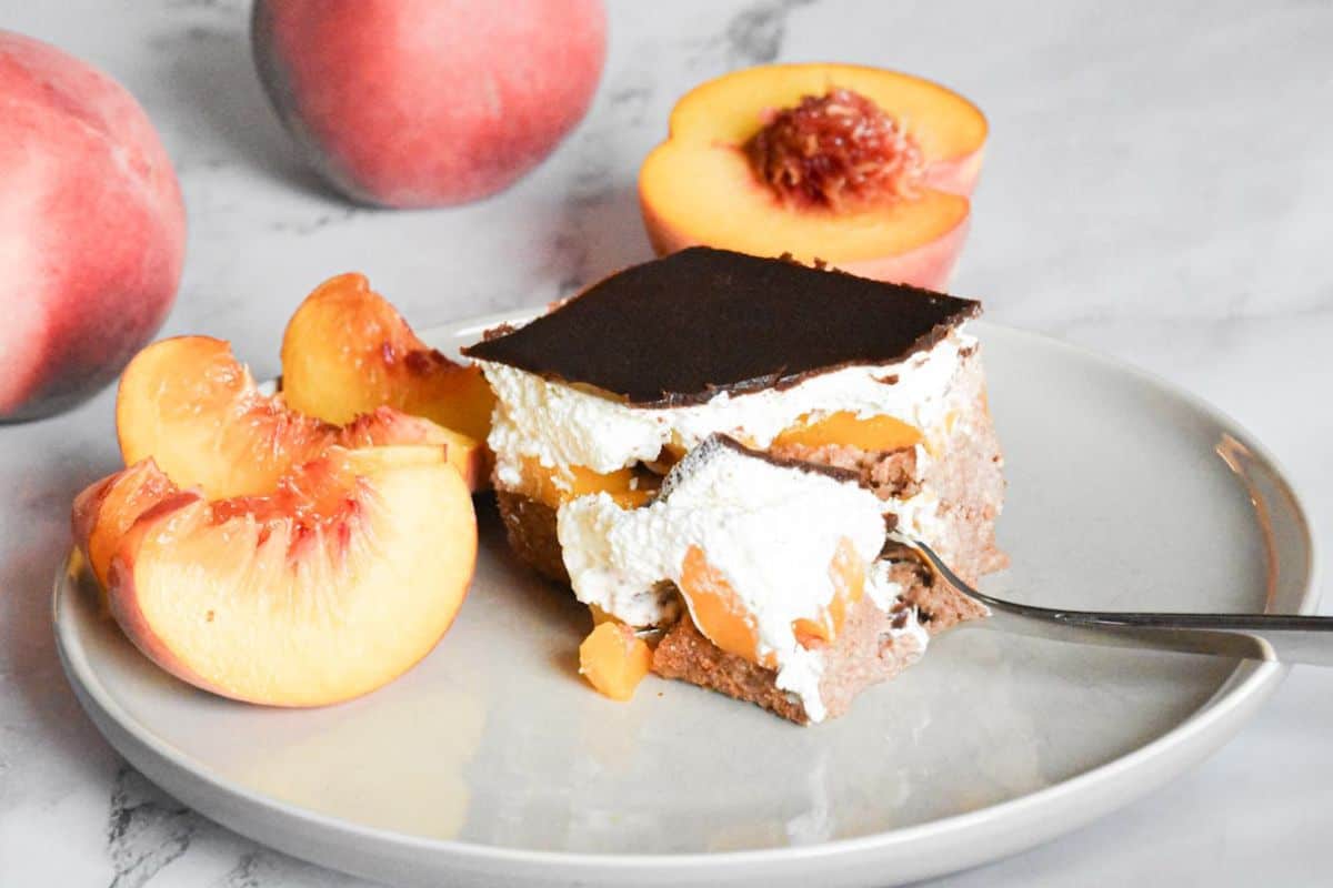 slice of Špikov Vrh cake on a plate being gently cut with a fork, revealing layers of chocolate sponge, cream, and glaze, with peach slices nearby.