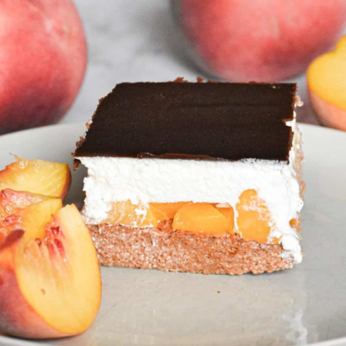 A slice of Špikov Vrh cake served on a plate, showing layers of chocolate sponge, cream, and glaze, with peach slices placed nearby.
