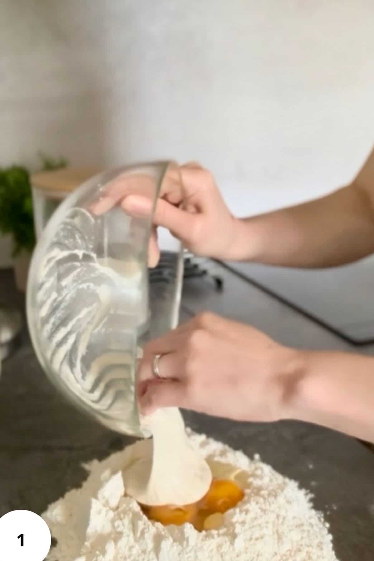 A person pouring flour and other ingredients from a bowl onto a clean surface to begin mixing the dough.