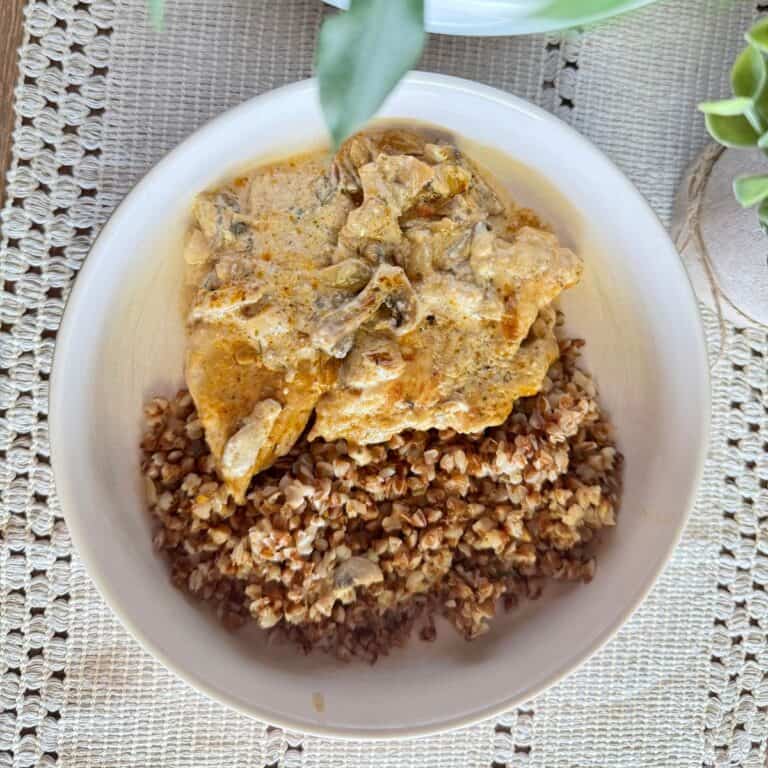 Close-up of creamy chicken with mushroom sauce served alongside buckwheat on a plate, with decorative plants on the side.