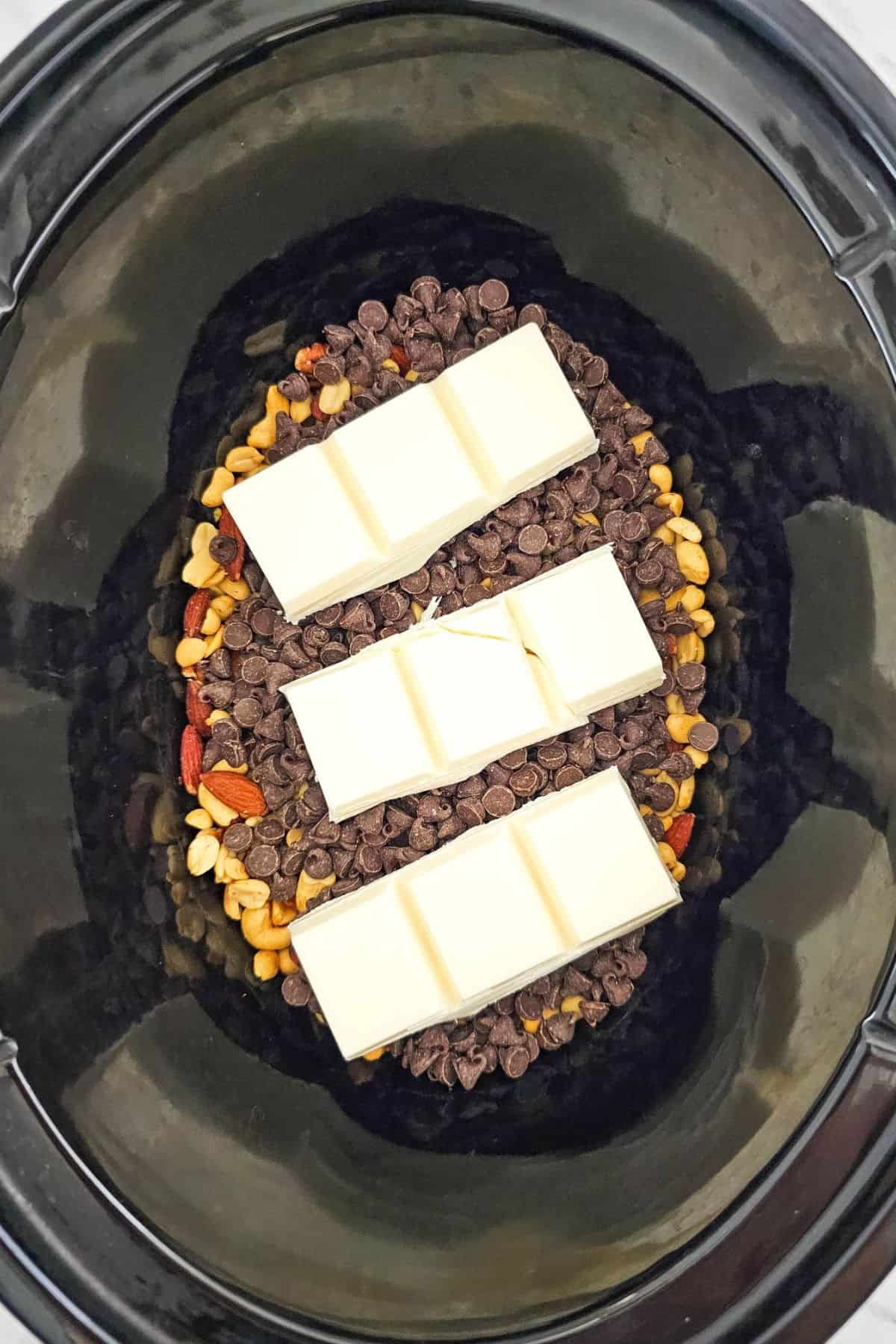 Ingredients for holiday crockpot candy including mixed nuts, chocolate chips, and white almond bark being placed in a Crockpot.