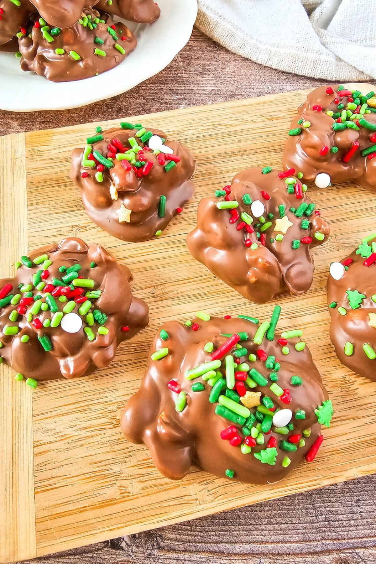 Crockpot holiday candy served on a wooden board with chocolate-coated and festive sprinkles.
