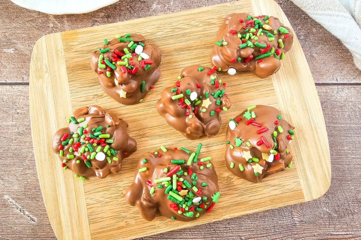 Crockpot holiday candy served on a wooden board with chocolate-coated and festive sprinkles.