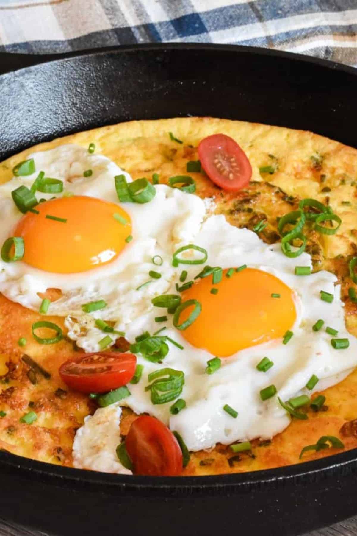 Dutch Baby pancake in a skillet, puffed and golden, topped with sunny-side-up eggs and garnished with fresh herbs.