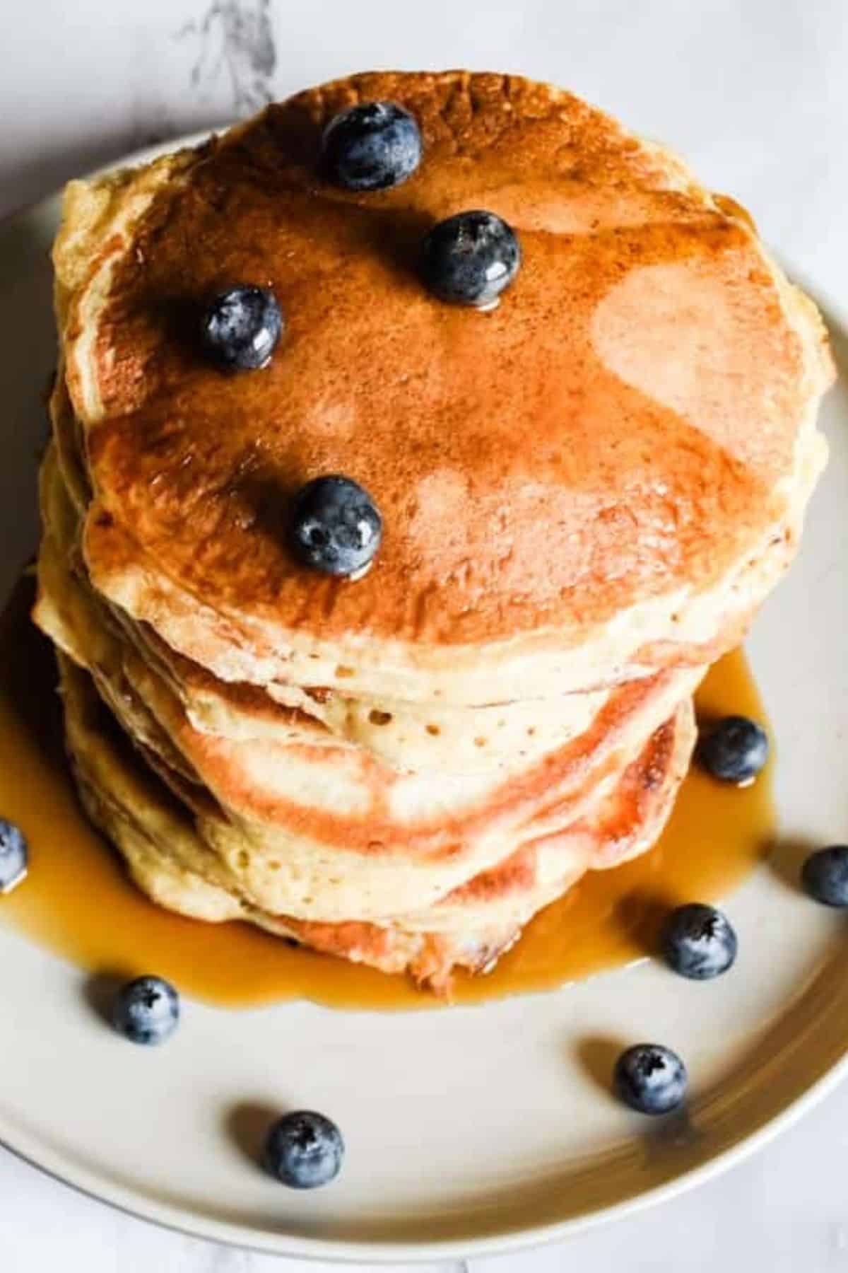 Stack of fluffy pancakes on a white plate, topped with fresh blueberries and syrup.