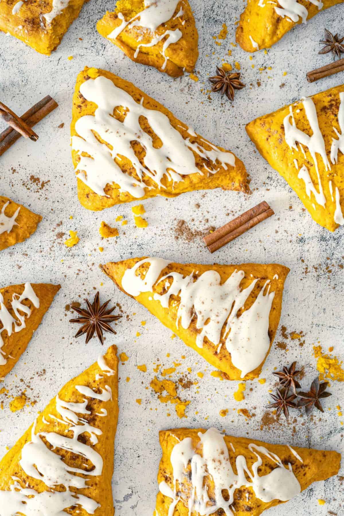 Spiced pumpkin scones with vanilla glaze on a surface, surrounded by cinnamon sticks.