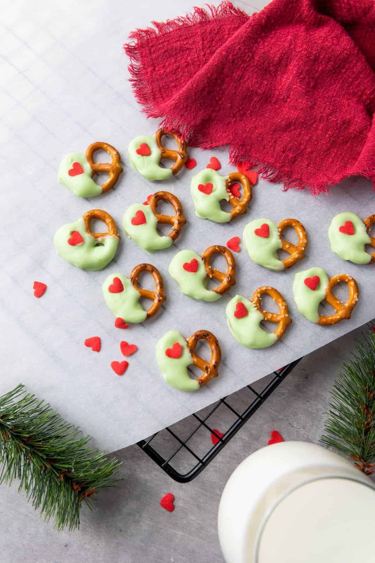 Christmas pretzels coated in green candy melts with red heart sprinkles, cooling on a wire rack lined with parchment paper, surrounded by additional sprinkles.