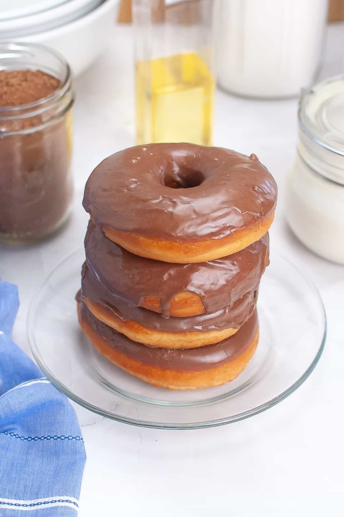 Stacked chocolate donuts on a plate with baking ingredients blurred in the background.