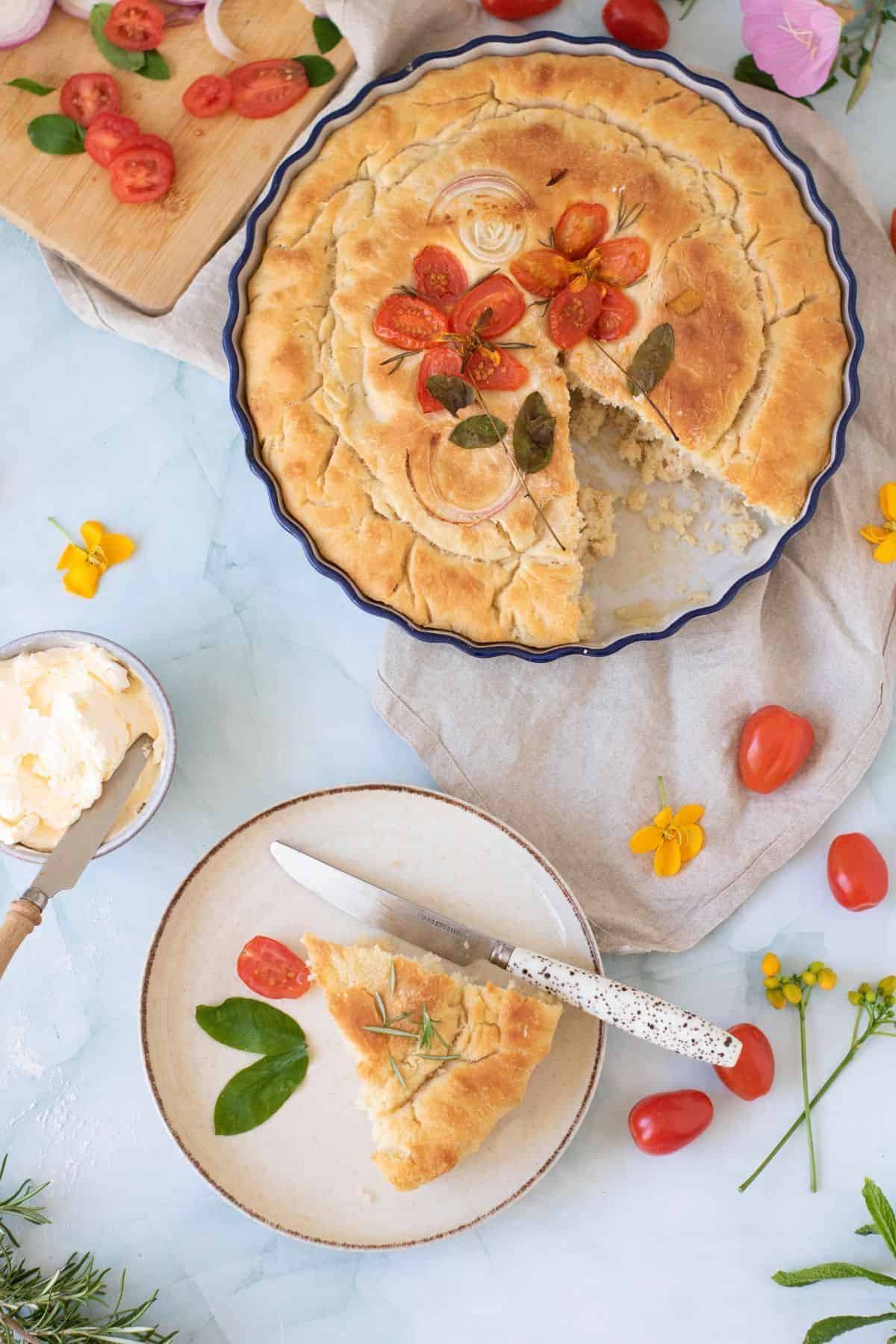 Focaccia with cherry tomatoes and onion served on a plate with a knife, with the whole focaccia placed on the side.