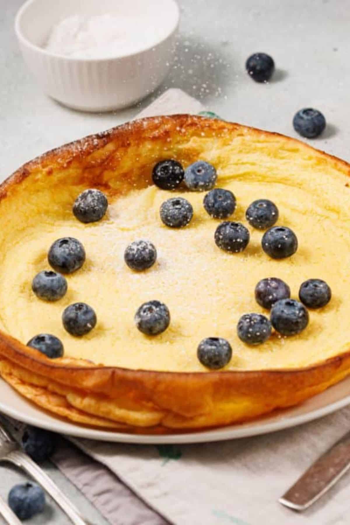 Dutch Baby Pancake dusted with powdered sugar and topped with berries, surrounded by fresh blueberries on the surface.