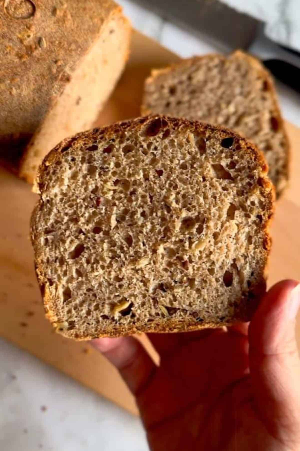 A hand holding a single rustic rye sourdough slice with the full loaf in the background.