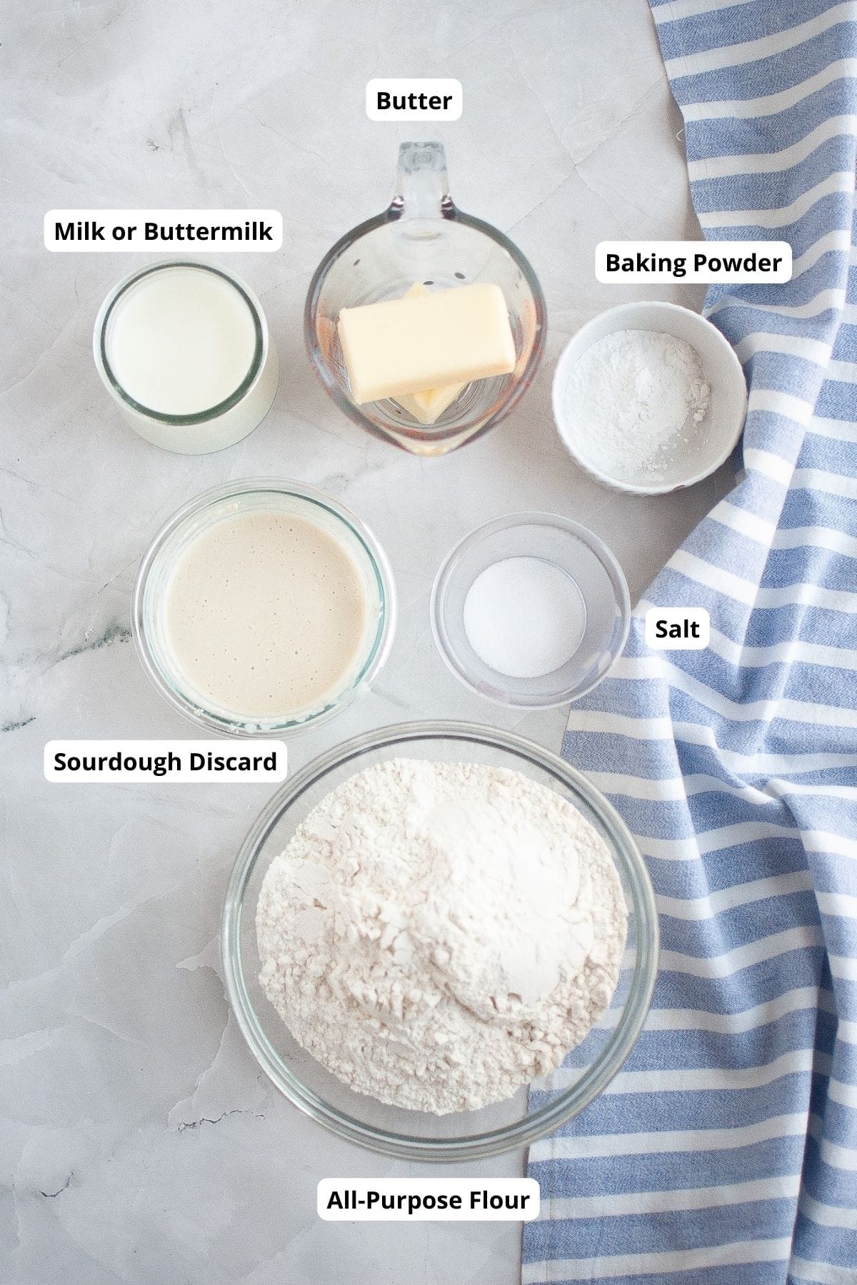 Ingredients for sourdough drop biscuits, including flour, sourdough discard, milk, butter, baking powder, and salt, arranged neatly on a surface with a striped cloth on the side.