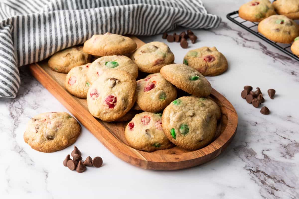 A stack of Christmas M&M cookies served on a wooden board with colorful candies visible with cloth underneath.