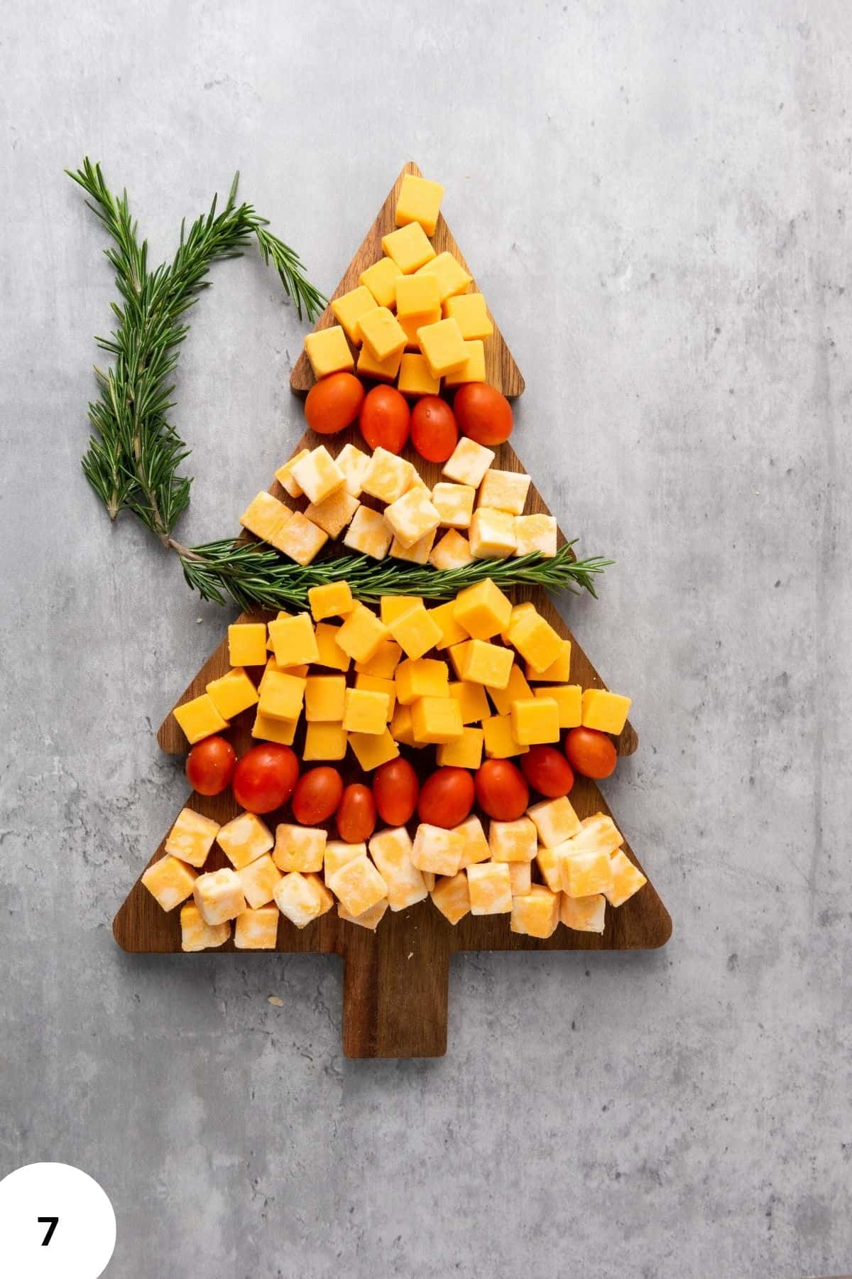 Christmas tree-shaped cheese platter with Colby Jack and Gouda cubes, cherry tomatoes, and rosemary, surrounded by festive decorations and cloth on the side.