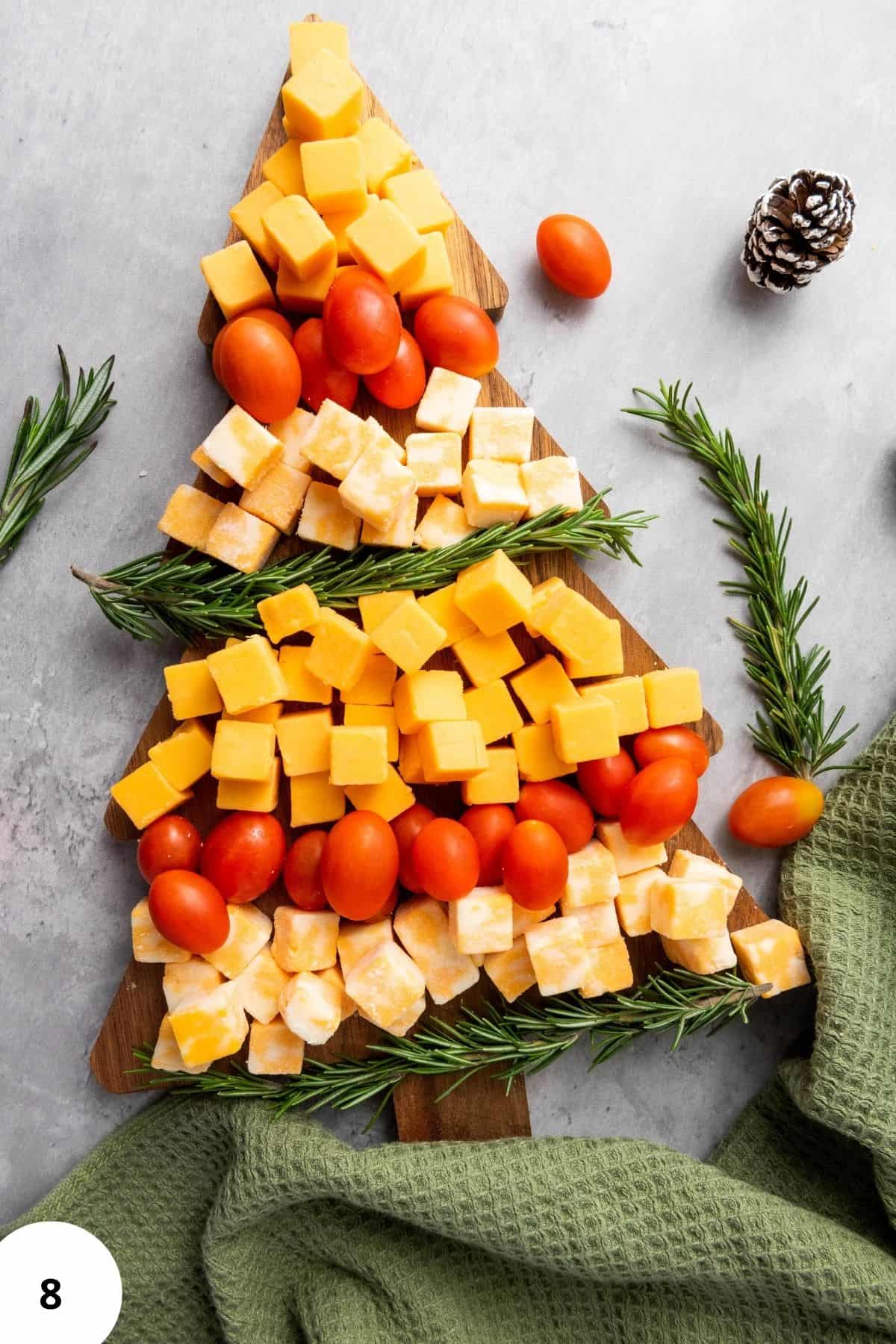 Christmas tree-shaped cheese platter with Colby Jack and Gouda cubes, cherry tomatoes, and rosemary, surrounded by festive decorations and cloth on the side.