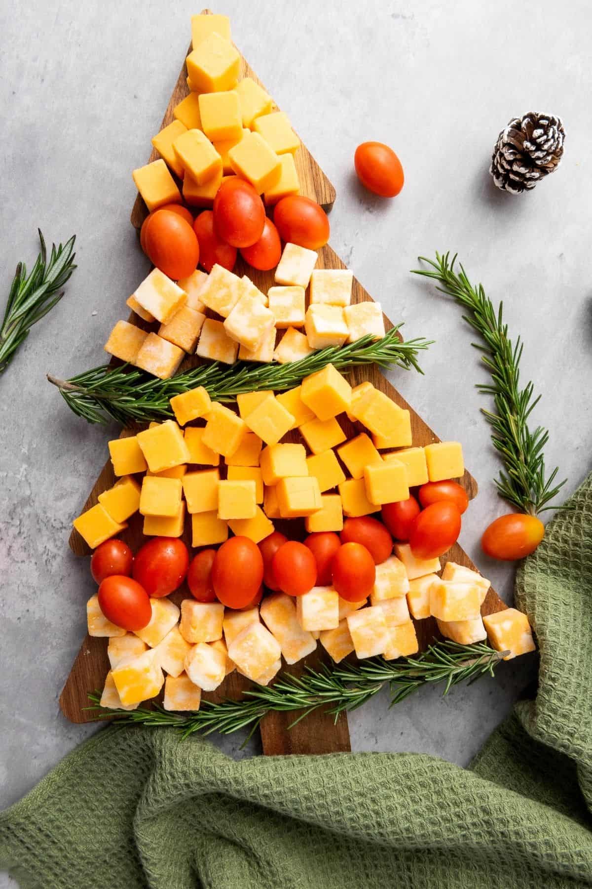 Christmas tree-shaped cheese platter with Colby Jack and Gouda cubes, cherry tomatoes, and rosemary, surrounded by festive decorations and cloth on the side.