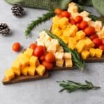 Christmas tree-shaped cheese platter with Colby Jack and Gouda cubes, cherry tomatoes, and rosemary, surrounded by festive decorations and cloth on the side.