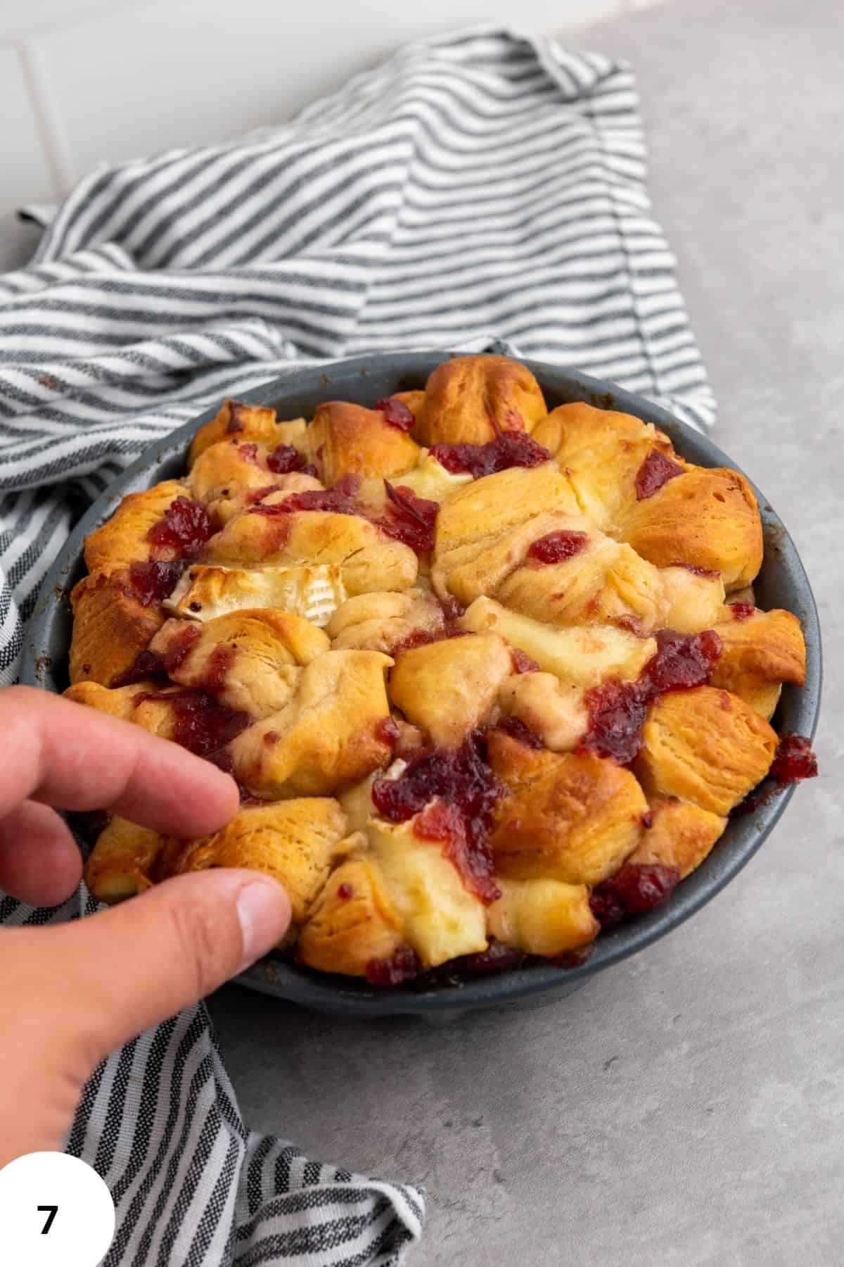 A hand holding a piece of bread with the rest of the loaf in the background.