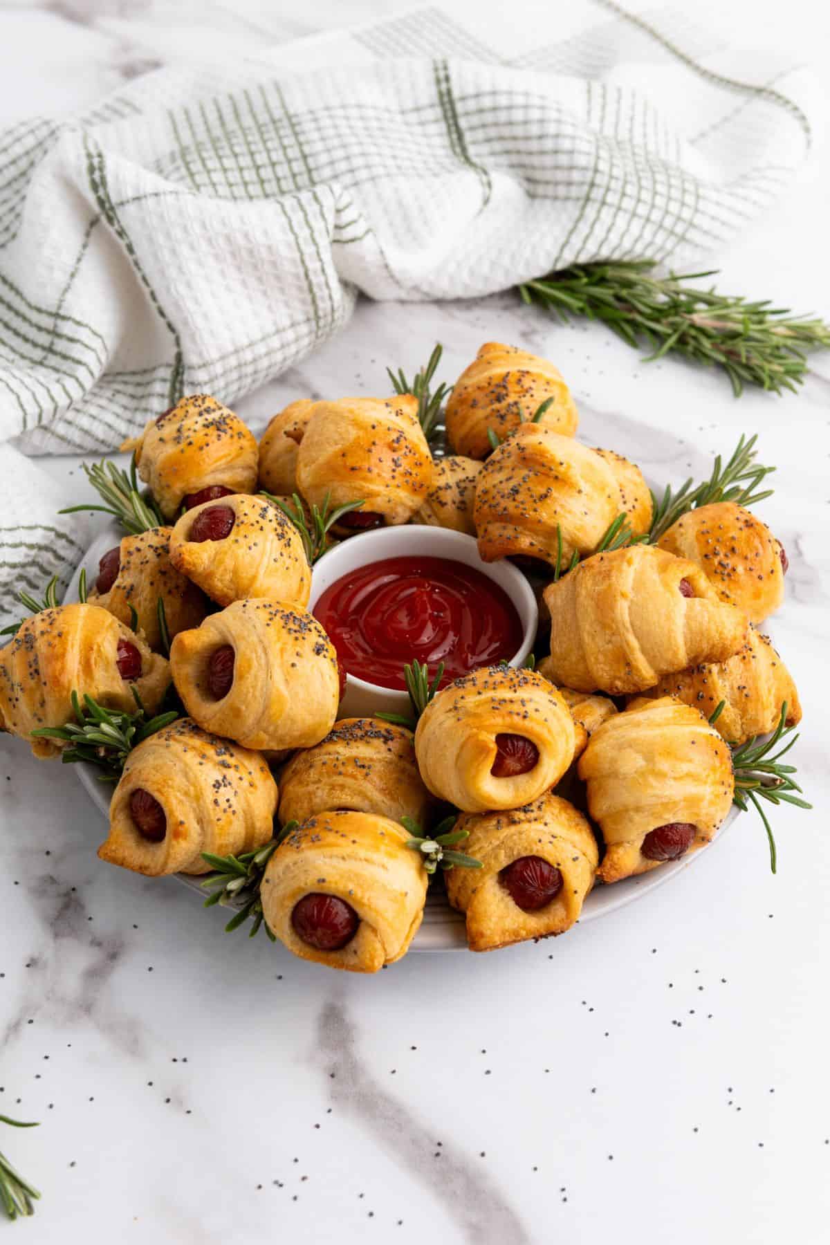 Mini piggies in croissant dough arranged on a plate, garnished with herbs, with a dipping sauce in the center.
