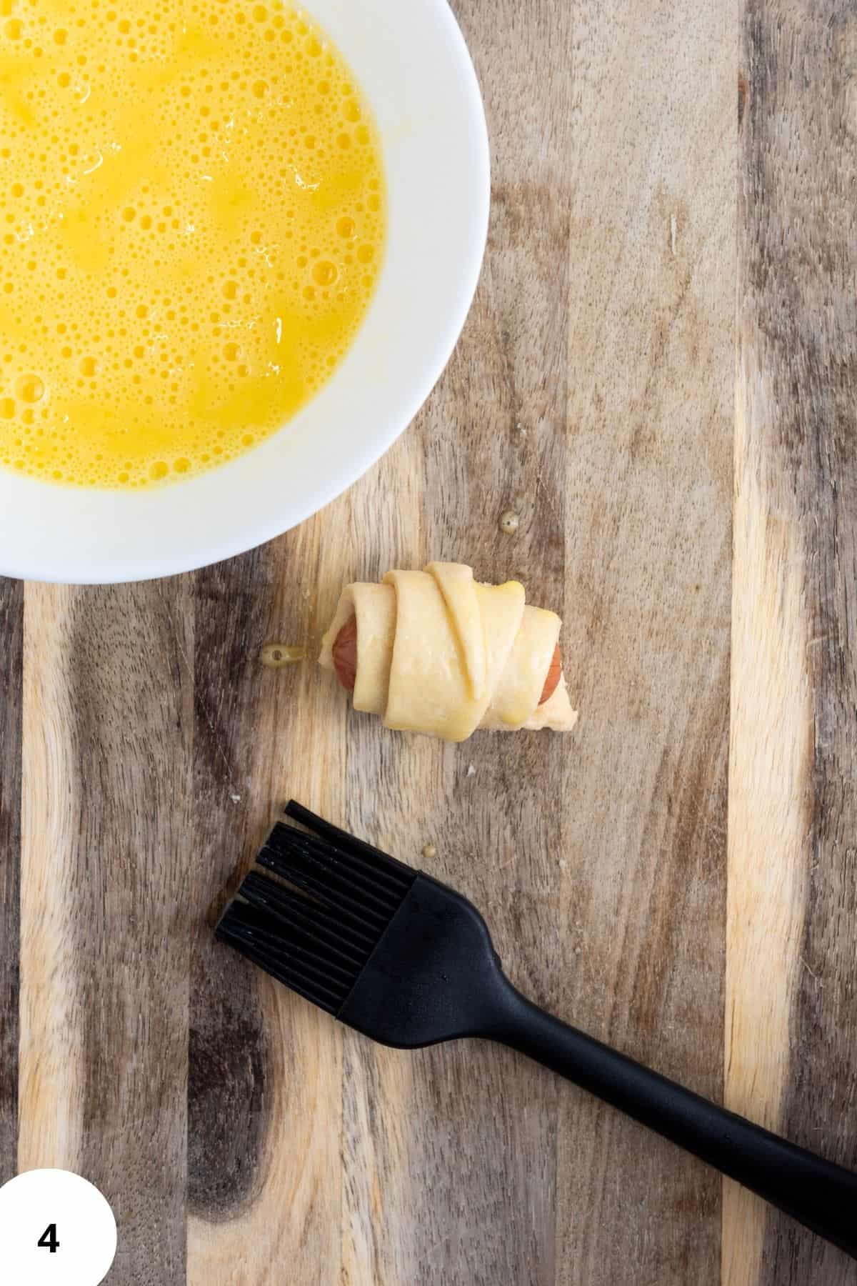 Rolled mini piggies in croissant dough on a wooden board, with a small bowl of egg wash and a brush on the side.