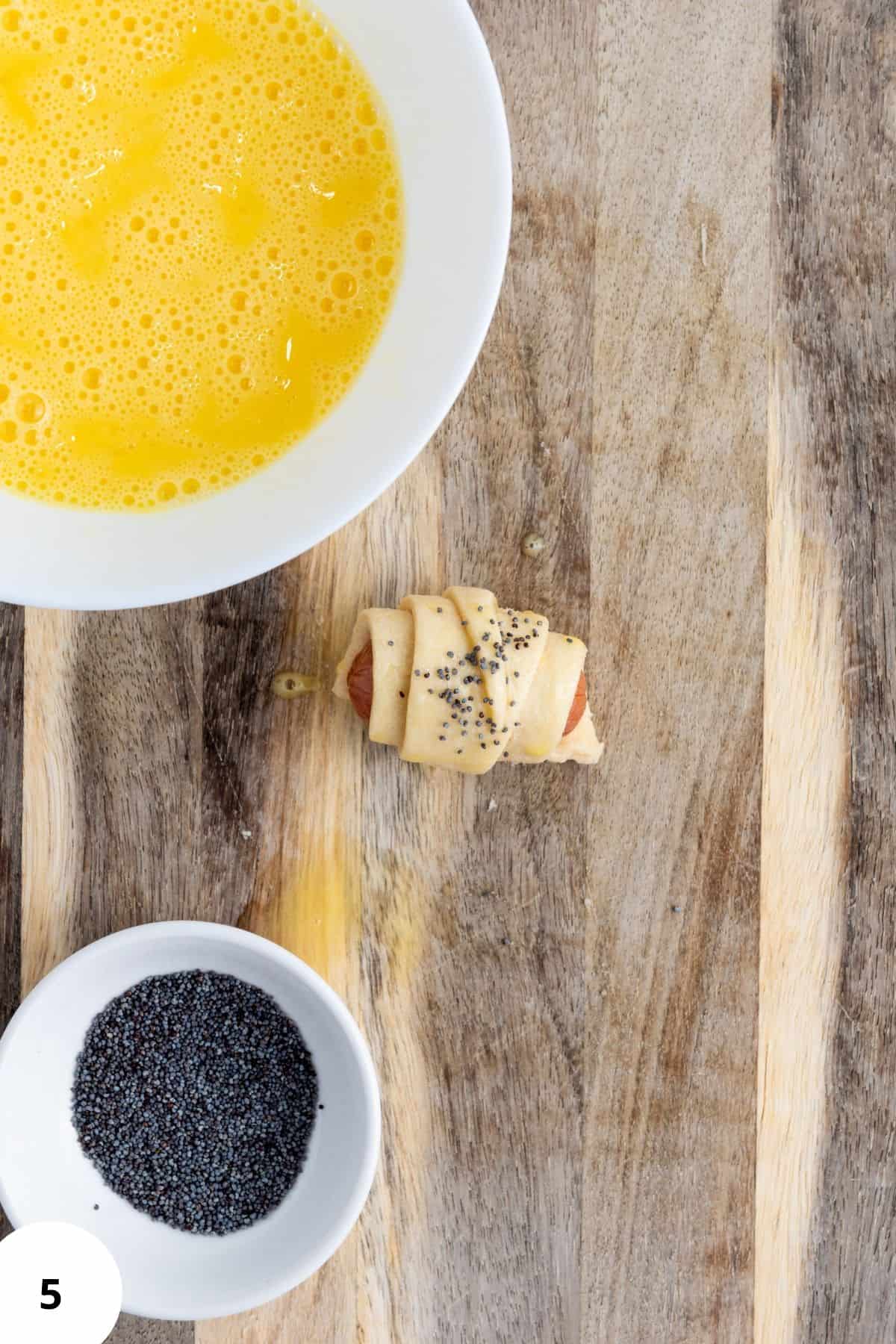 Rolled mini piggies in croissant dough on a wooden board, topped with poppy seeds.