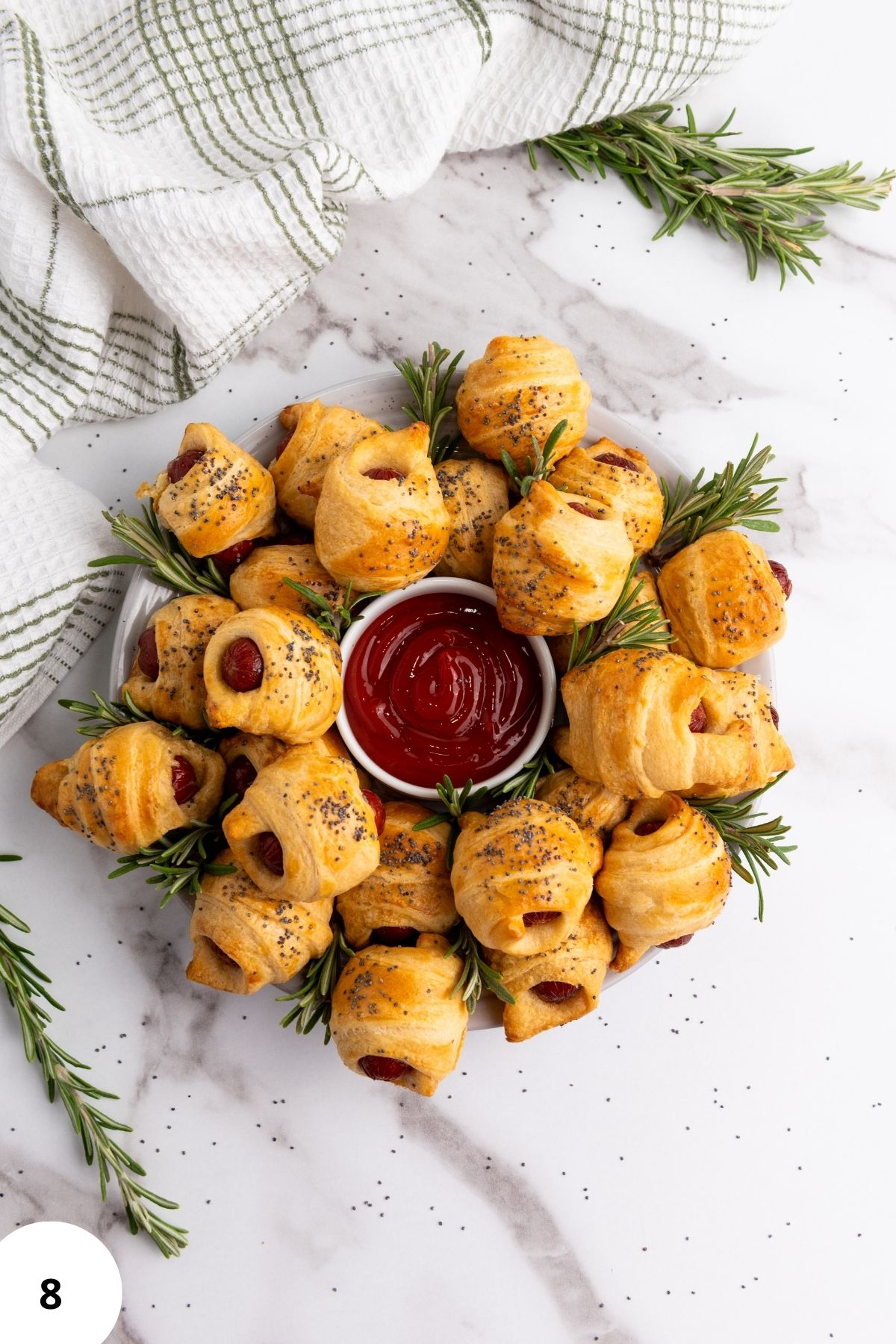 Mini piggies in croissant dough arranged on a plate, garnished with herbs, with a dipping sauce in the center.