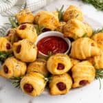 Mini piggies in croissant dough arranged on a plate, garnished with herbs, with a dipping sauce in the center.