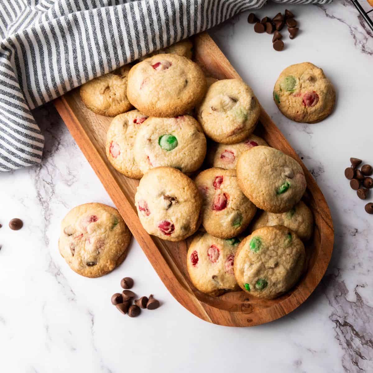 A stack of Christmas M&M cookies served on a wooden board with colorful candies visible with cloth underneath.