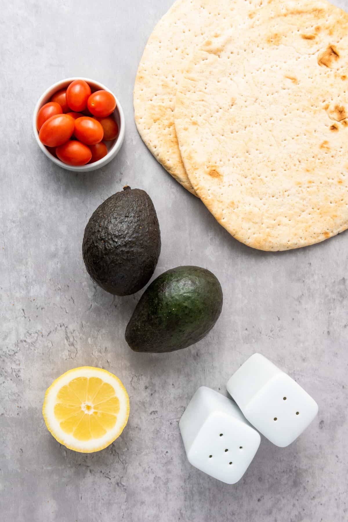 All ingredients for avocado pita tree appetizers arranged on a surface, including pita bread, avocados, cherry tomatoes, garlic, lemon juice, salt, and pepper.