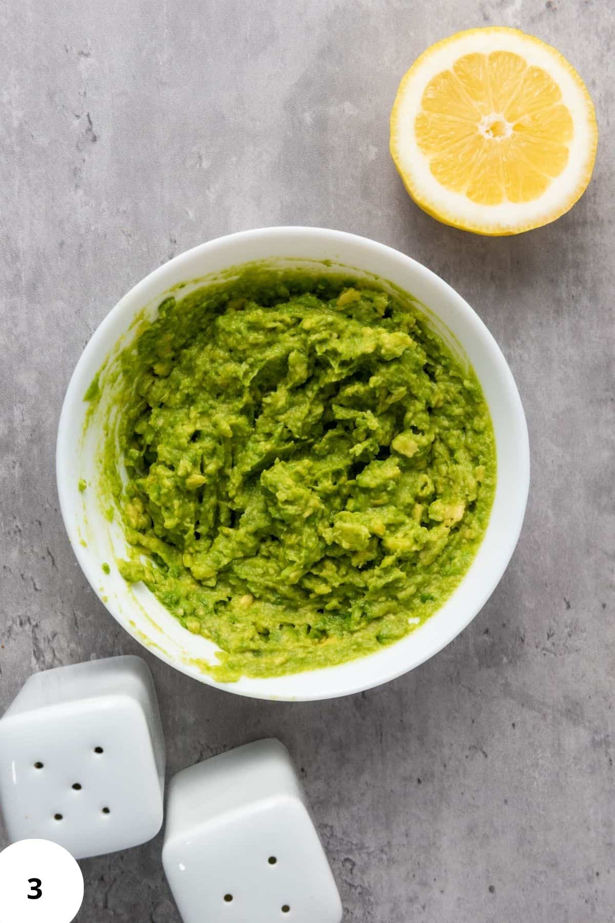 Mashed avocado in a bowl with a fork resting inside, surrounded by lemon and seasonings.