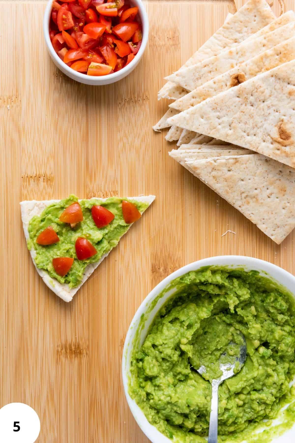 A single pita wedge being spread with creamy mashed avocado, topped with cherry tomatoes and surrounded with ingredients.