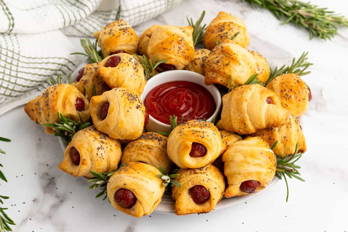 Mini piggies in croissant dough arranged on a plate, garnished with herbs, with a dipping sauce in the center.