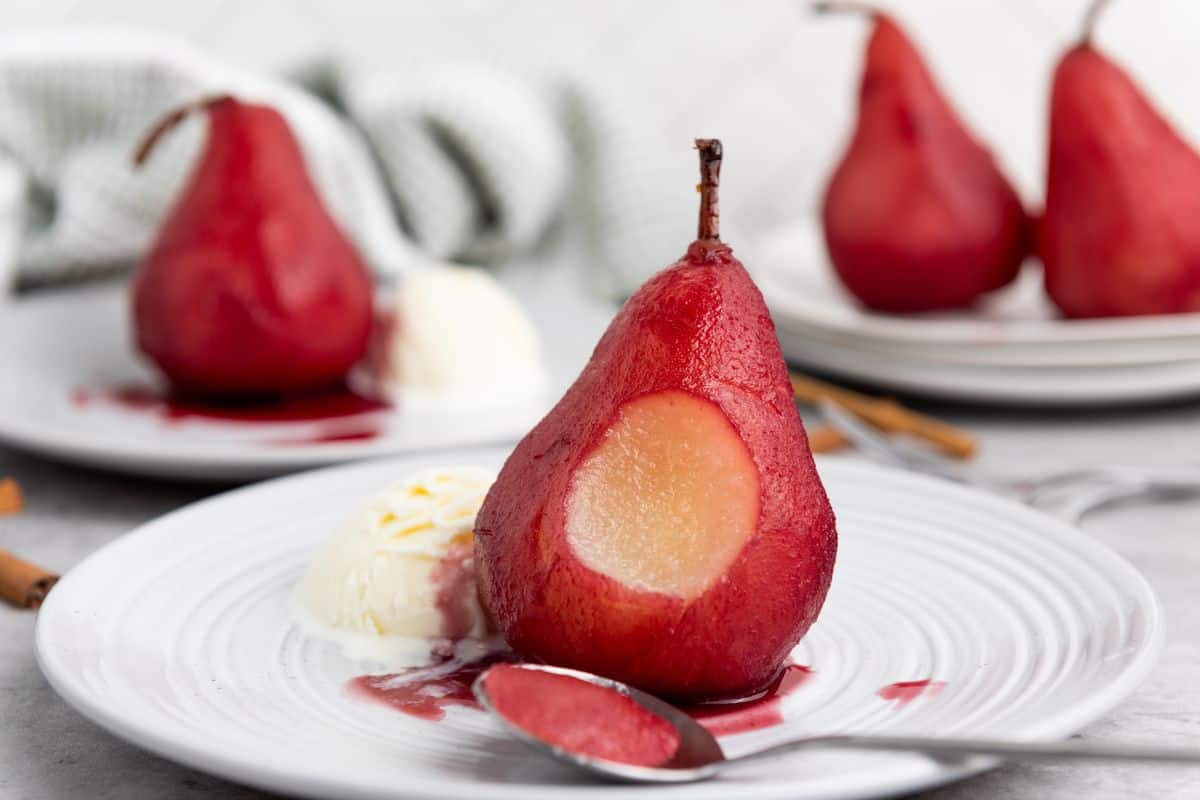 A plate with a single red wine poached pear bitten in the center, served with a scoop of vanilla ice cream on the side and a spoon beside it.