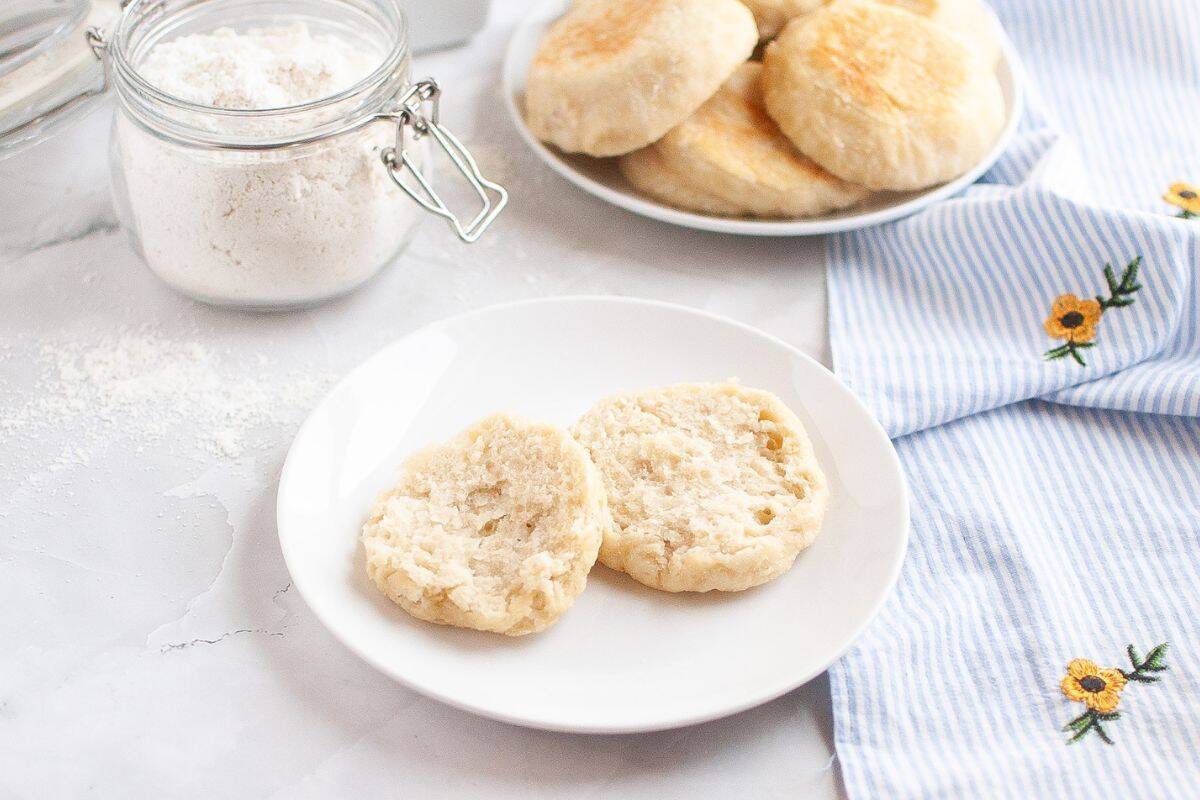 Sliced sourdough English muffins served on a plate in the foreground, with a jar of flour and more muffins on a plate softly blurred in the background.