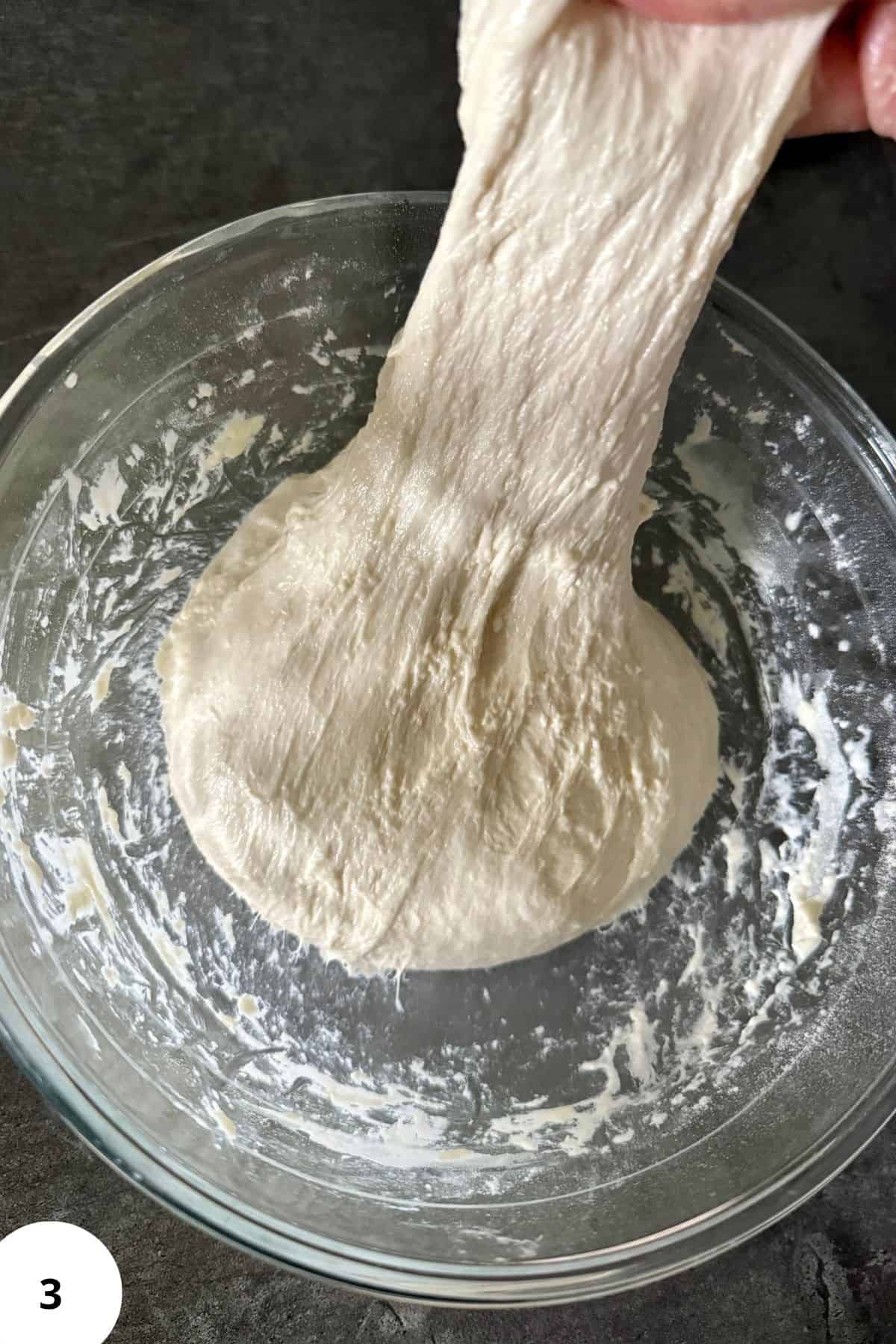 Dough in a mixing bowl being gently stretched by hand.