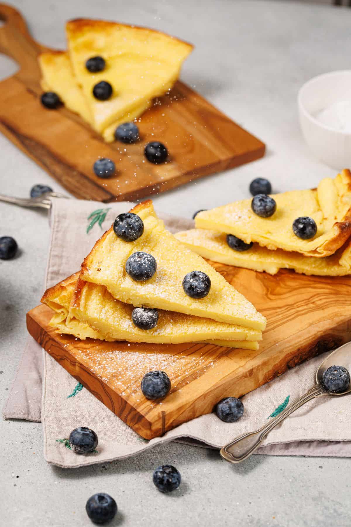 Sliced Dutch Baby pancake dusted with powdered sugar, served on a wooden board with fresh blueberries.