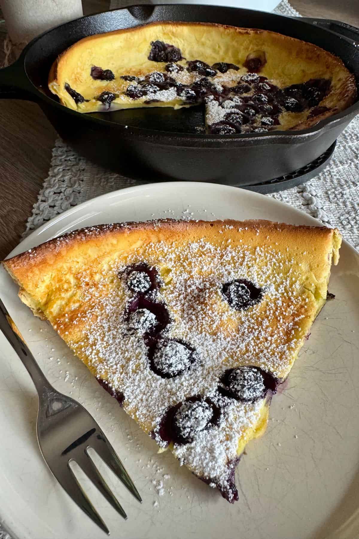 Single slice of blueberry Dutch Baby pancake served on a plate topped with fresh blueberries and powdered sugar.