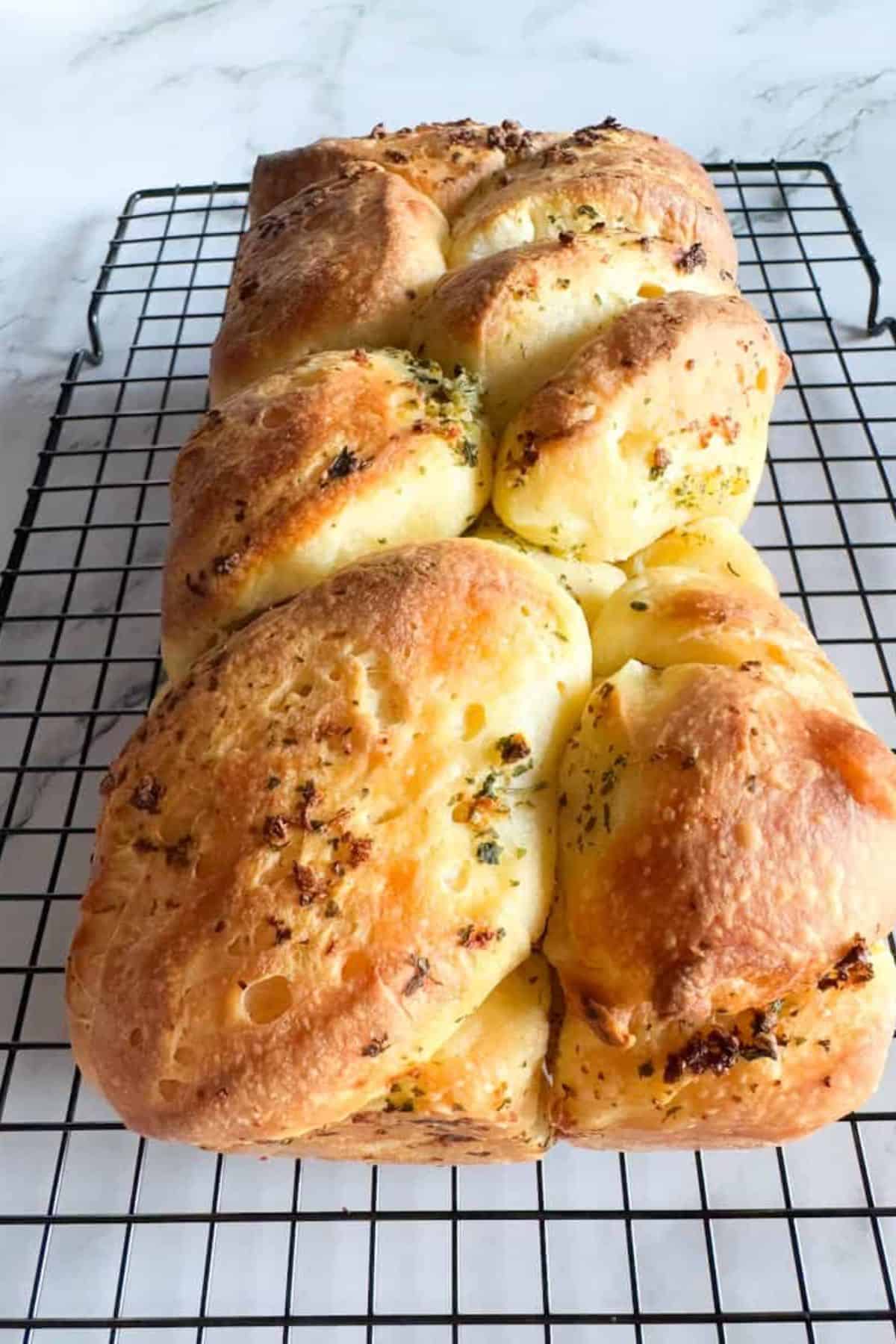 Golden-brown bread rolls with herbs on top, fresh out of the oven on a cooling rack, showcasing skillful dough stretching and folding.