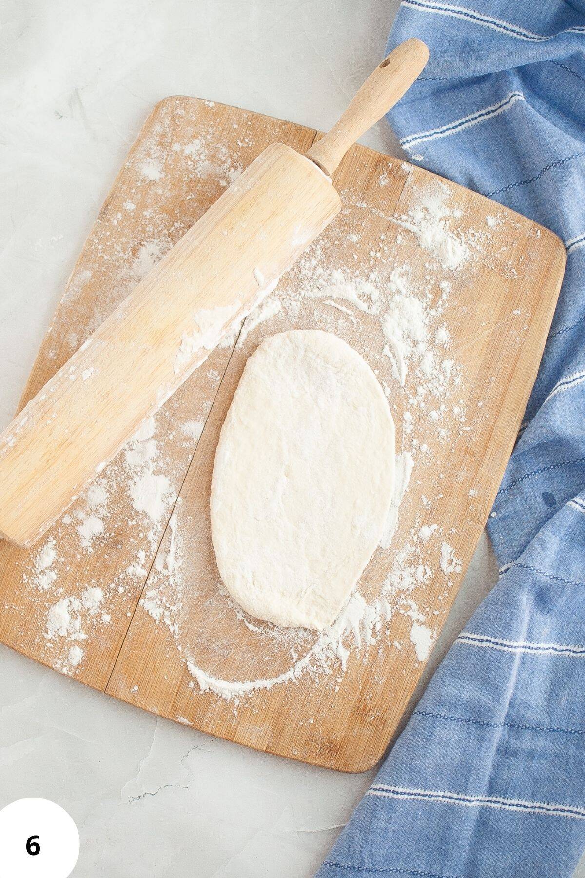 Flattened dough on a floured wooden board with a rolling pin and a tablecloth on the side.